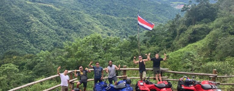 Group of travelers posing with ATVs at a scenic mountain viewpoint near Jaco, Costa Rica, with lush green hills and a Costa Rican flag.