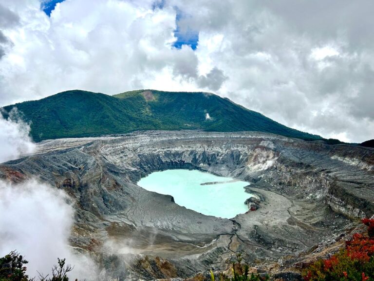 Turquoise crater lake at Poas Volcano in Costa Rica, surrounded by volcanic rock, green mountains, and clouds.
