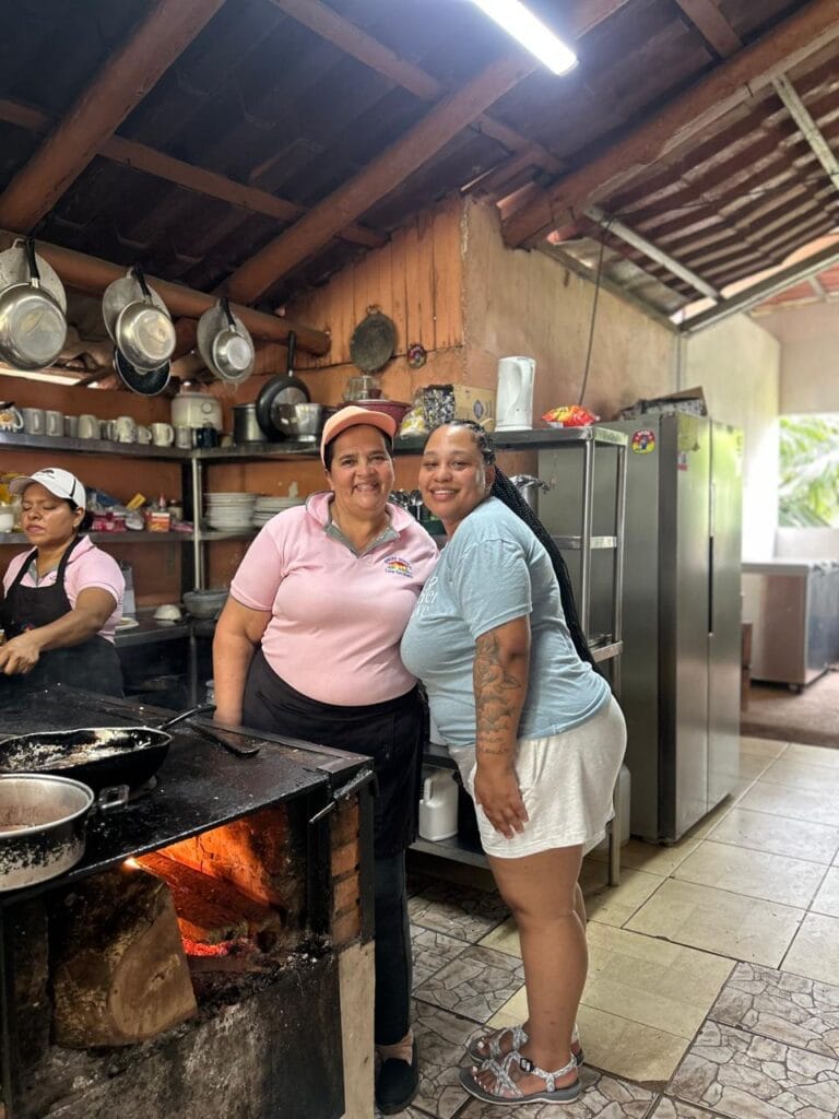 Doña Marta standing beside a happy guest in her rustic, wood-fired kitchen in the mountains near Jaco, Costa Rica. Traditional pots hang behind them, and a fire burns beneath the stove.